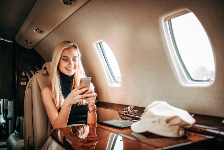 GoGo Wifi Young rich blonde woman while traveling on a private airplane. She is using a mobile phone. A hat and a digital tablet are on the table in front of her.